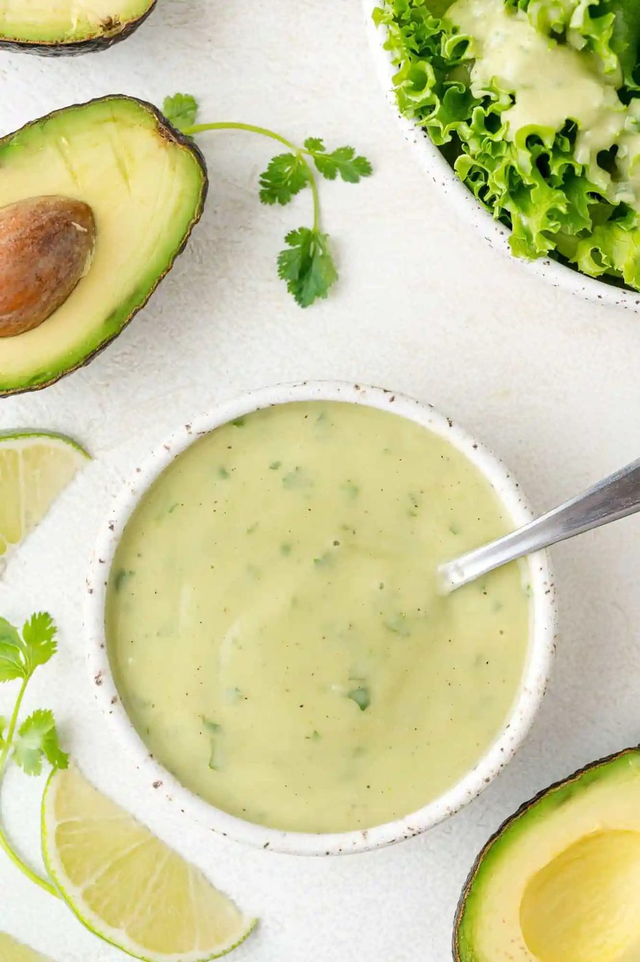 A bowl of creamy green dressing with herbs, surrounded by halved avocado, lime slices, and lettuce.