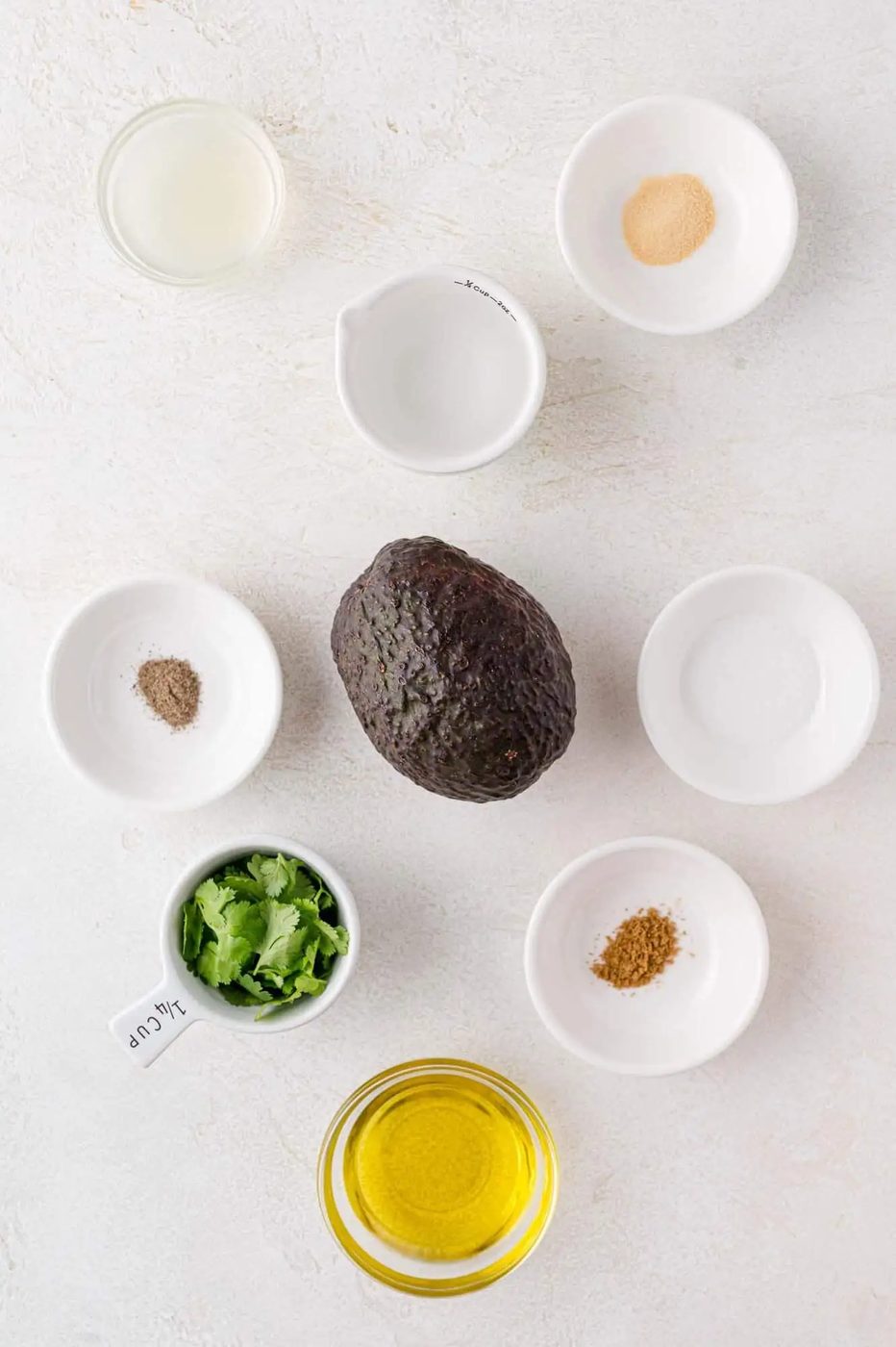 Ingredients for avocado dressing arranged on a surface: a whole avocado, olive oil, cilantro, lime juice, and seasonings in small bowls.