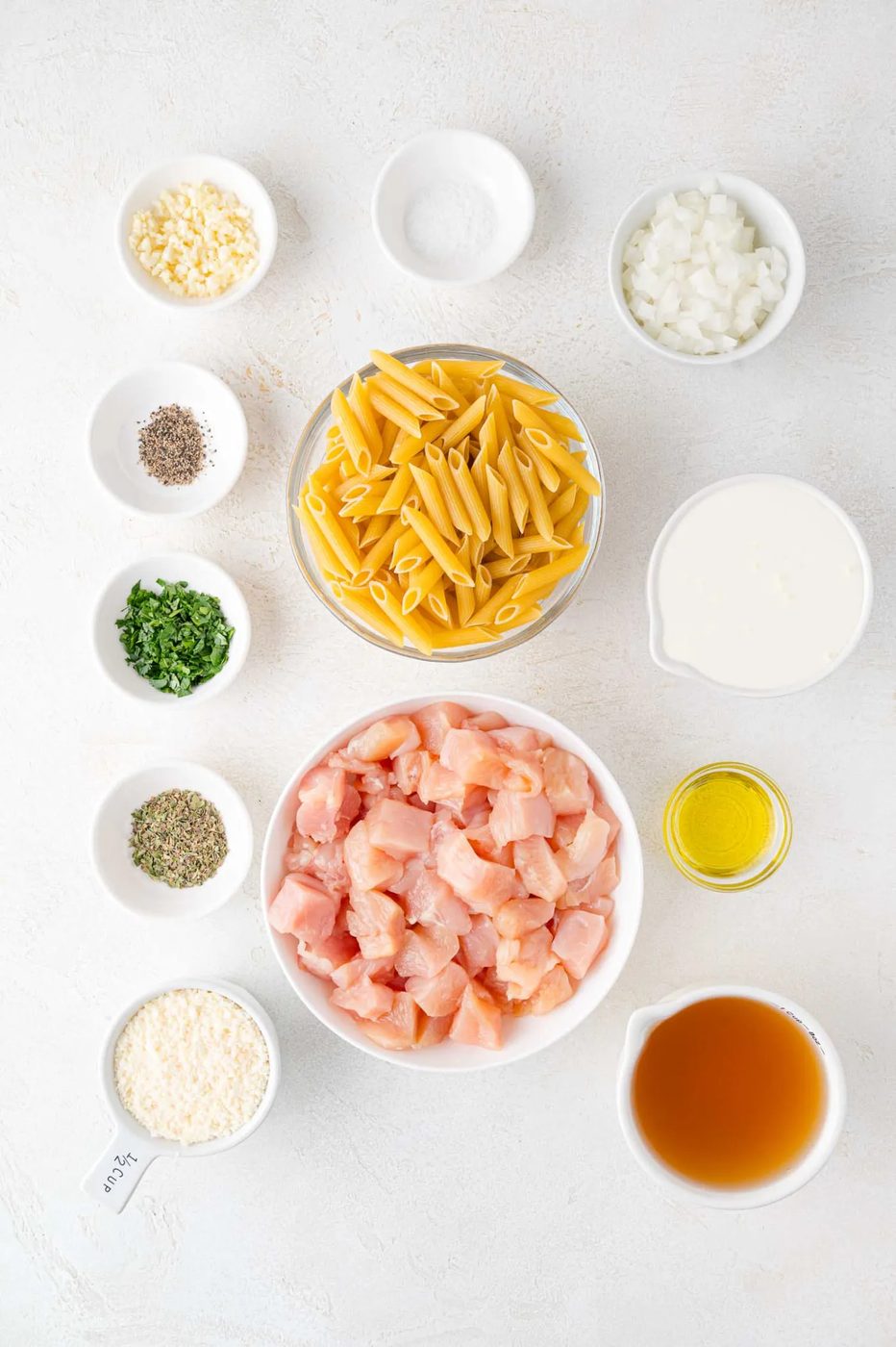 Ingredients for pasta dish include uncooked penne, diced chicken, diced onions, minced garlic, cream, broth, olive oil, grated cheese, and herbs, all arranged in bowls on a white surface.