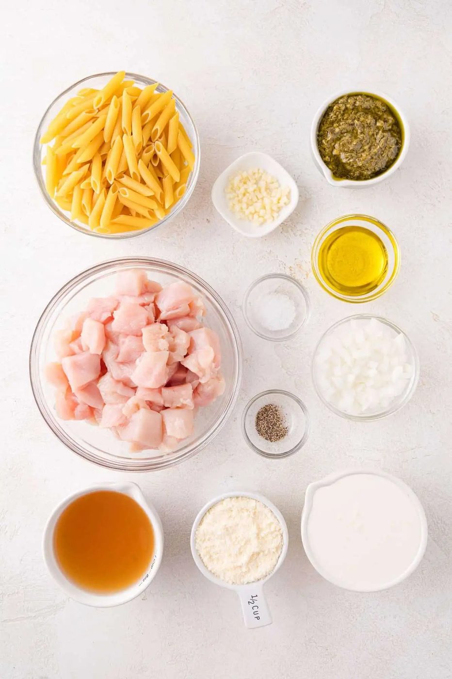 Ingredients for a pasta dish arranged on a white surface, including penne, diced raw chicken, pesto, olive oil, cream, grated cheese, chopped onion, minced garlic, broth, salt, and pepper.