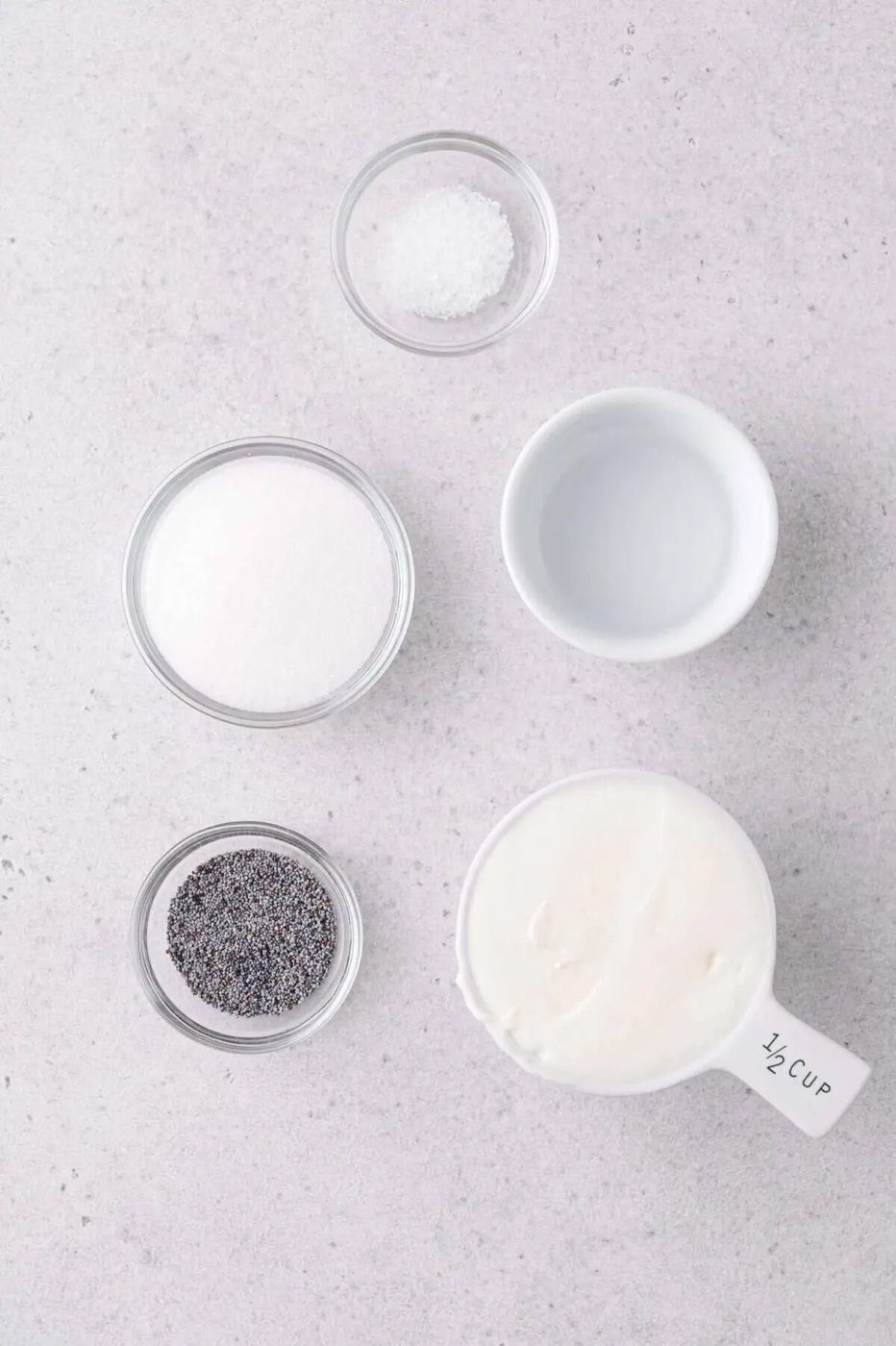 Five small bowls on a light countertop containing salt, sugar, poppy seeds, white vinegar, and mayonnaise.