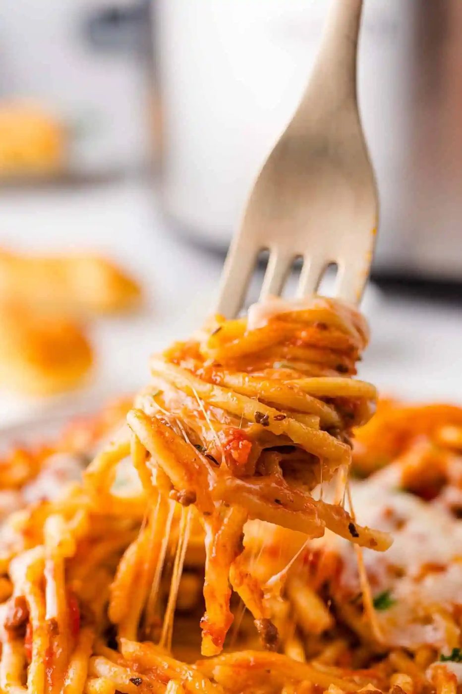 Fork lifting a portion of spaghetti with tomato sauce, cheese, and herbs, with cheese strands stretching from the plate.