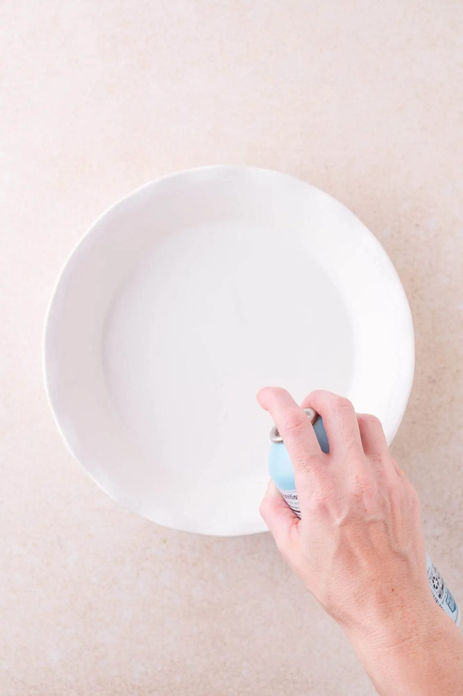 A hand sprays nonstick cooking spray into a round, white ceramic pie dish on a beige surface.