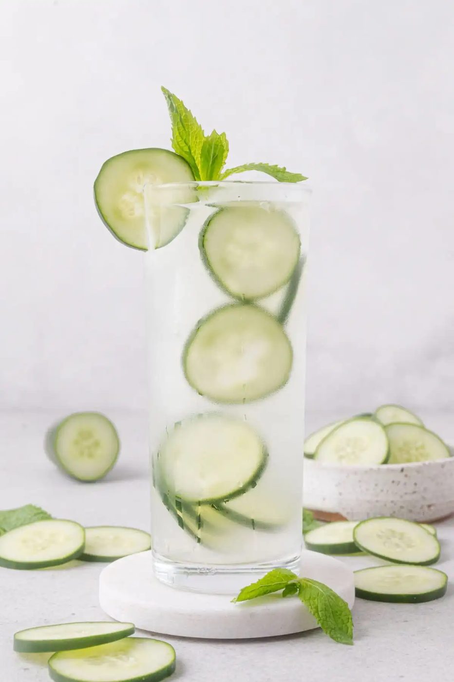A tall glass of cucumber lime cooler with ice, cucumber slices, and a sprig of mint, surrounded by cucumber slices on a light surface.