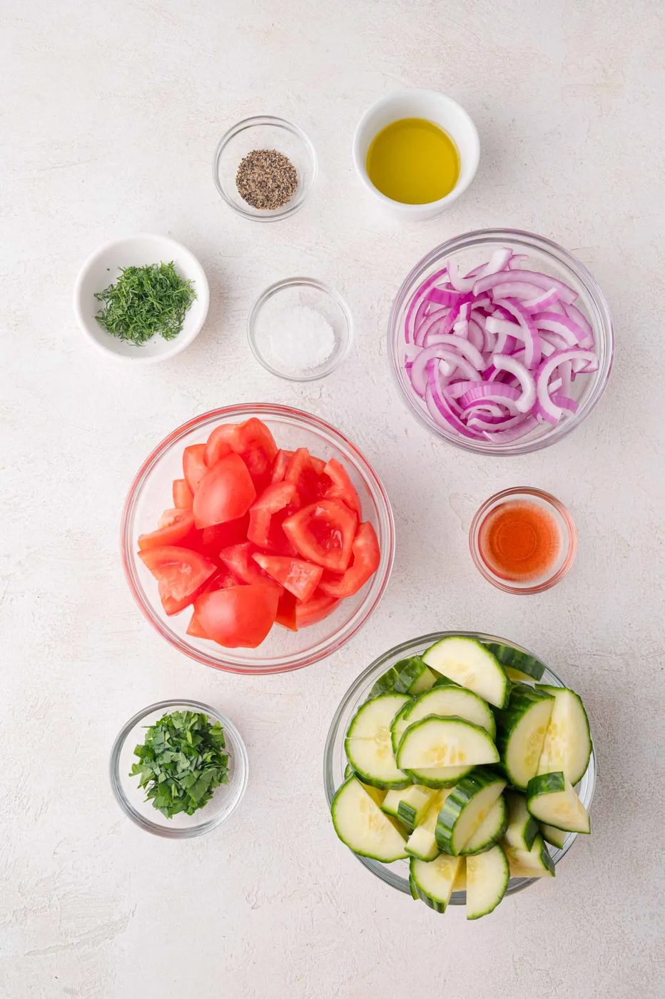Bowls of chopped tomatoes, sliced cucumbers, sliced red onions, chopped herbs, and small bowls of olive oil, vinegar, salt, and pepper arranged on a white surface.