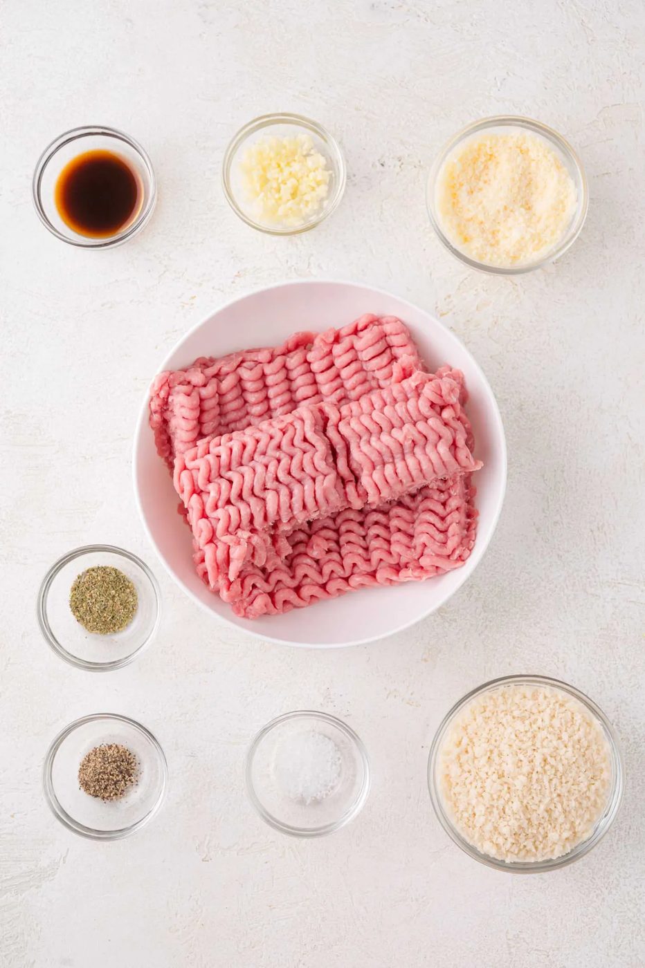 Raw ground turkey in a bowl surrounded by small bowls of grated cheese, breadcrumbs, minced garlic, black pepper, salt, and a dark liquid seasoning on a light surface.