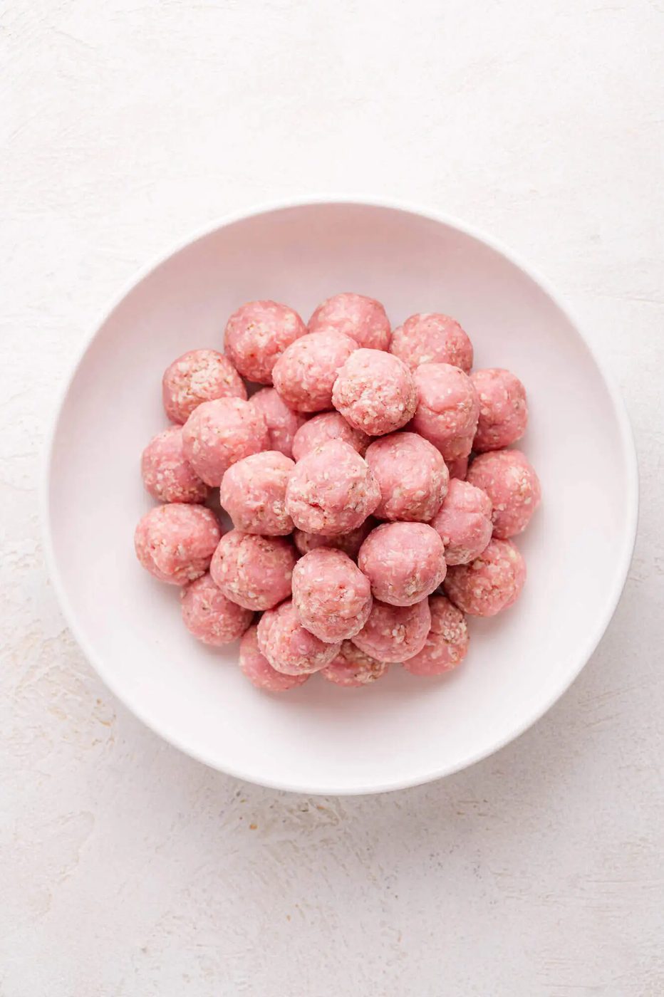 A white bowl filled with raw, uncooked meatballs made from ground turkey and seasoning, placed on a light surface.