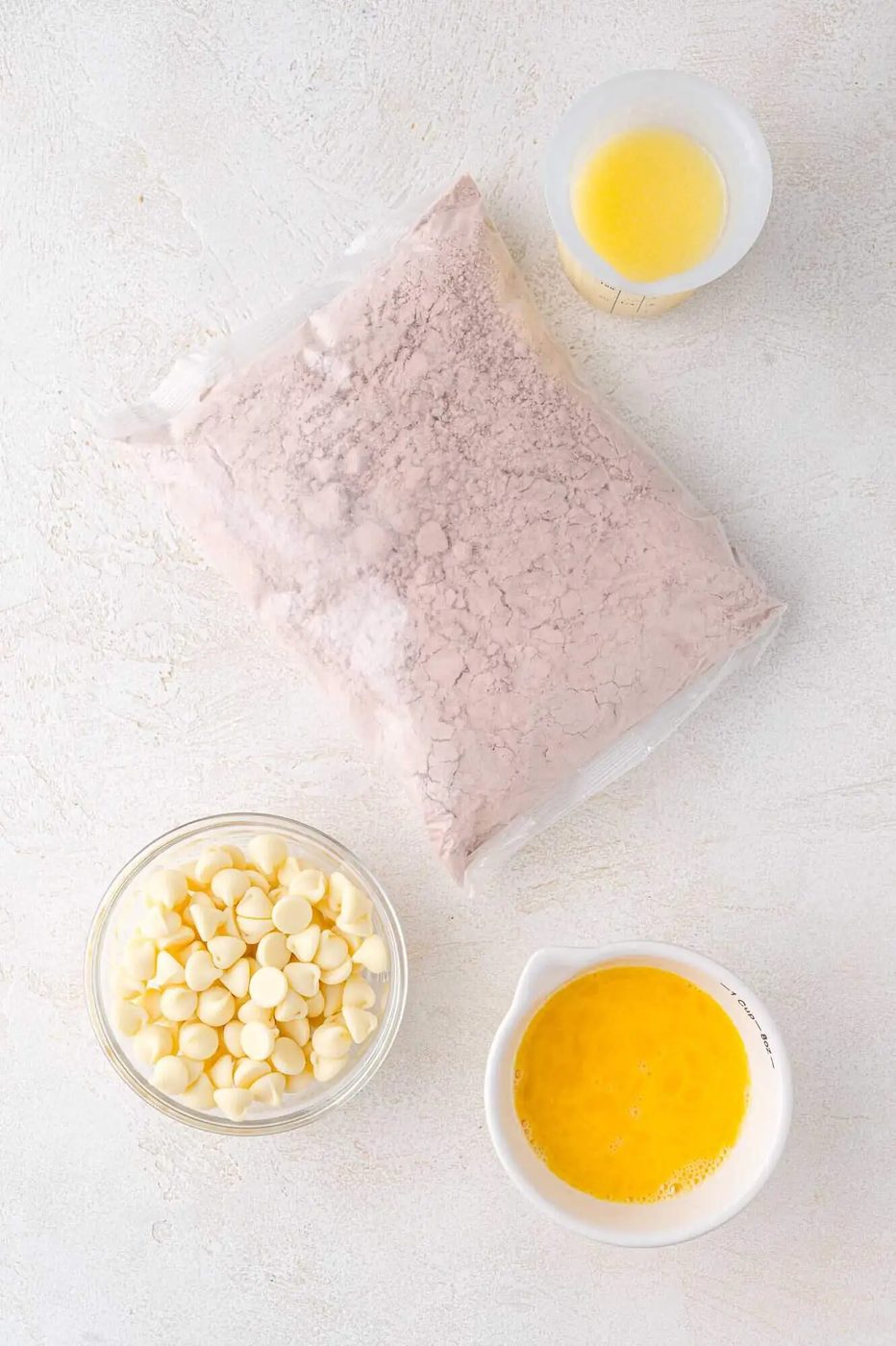A bag of red velvet cake mix, a bowl of white chocolate chips, a small bowl of melted butter, and a cup of beaten eggs arranged on a white surface.
