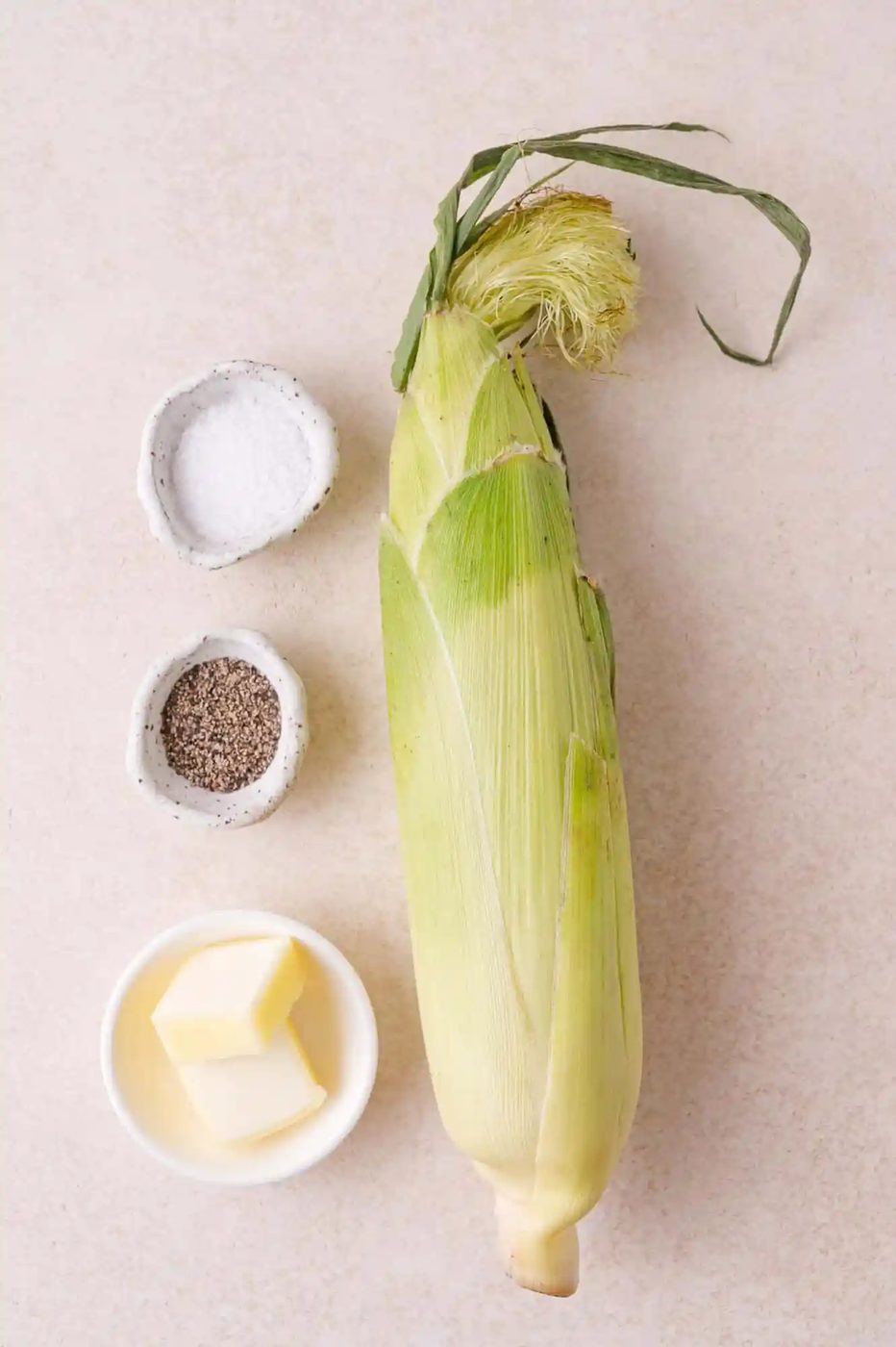 An ear of corn in husk, a small bowl of butter, and small bowls of salt and pepper are arranged on a light surface.
