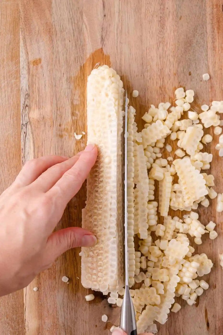 A hand holds a corn cob on a wooden surface while a knife slices off kernels, which are scattered nearby.