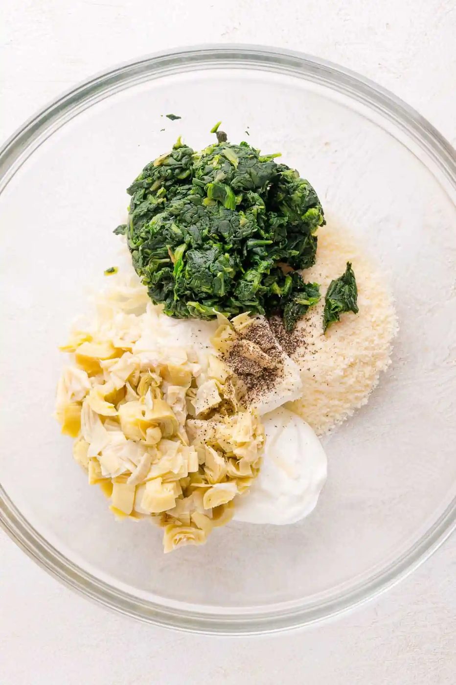 A glass bowl containing chopped spinach, artichoke hearts, grated cheese, mayonnaise, and seasoning on a white surface.