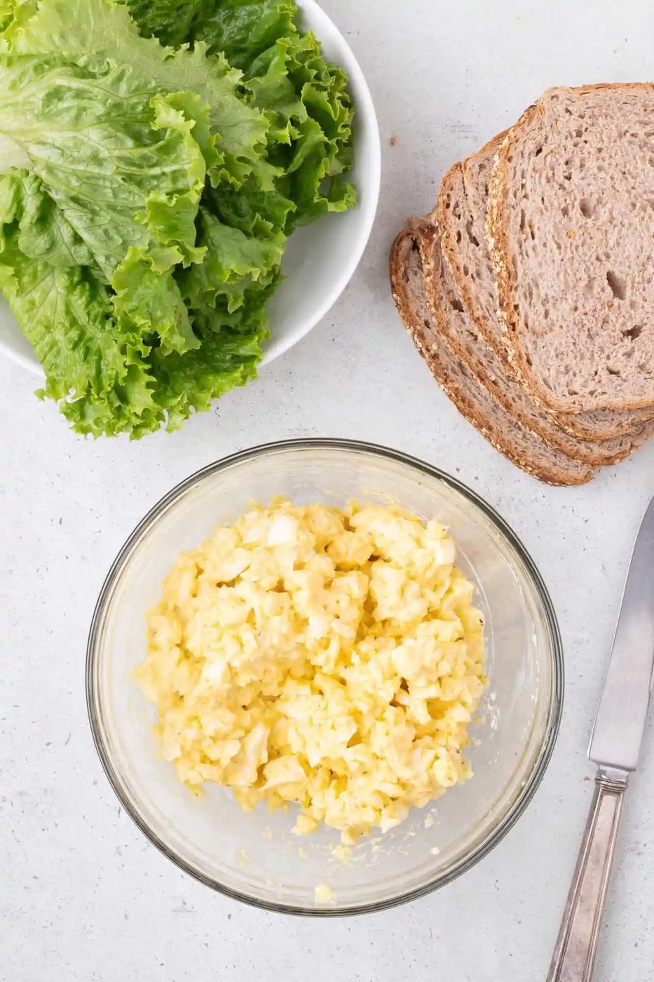 A bowl of egg salad next to a plate of lettuce, slices of brown bread, and a knife on a white surface.