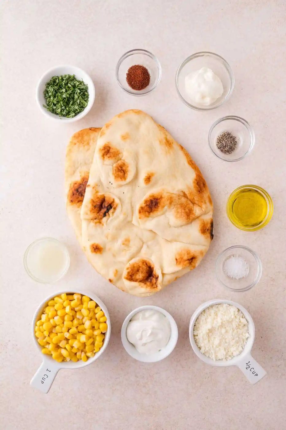 Assorted ingredients for a flatbread dish, including naan, corn, grated cheese, herbs, yogurt, mayonnaise, olive oil, spices, salt, pepper, and lemon juice in small bowls.