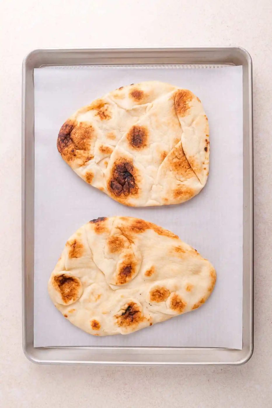 Two pieces of baked naan bread on a parchment-lined metal baking sheet.