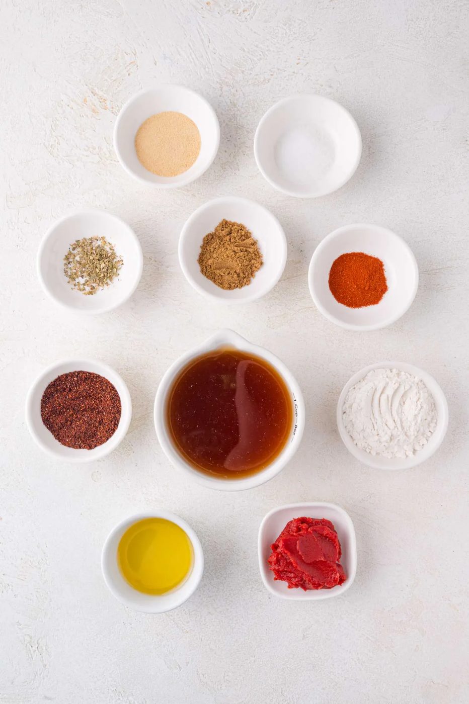 A variety of spices and ingredients are arranged in small white bowls on a light surface.