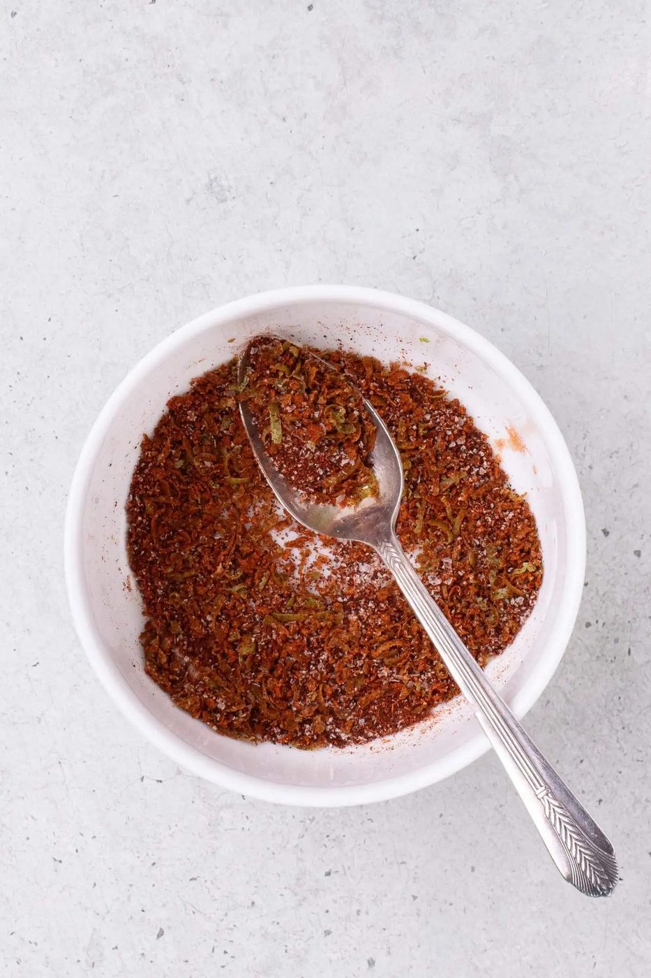 A bowl containing a mixture of red spices and herbs with a spoon resting inside.
