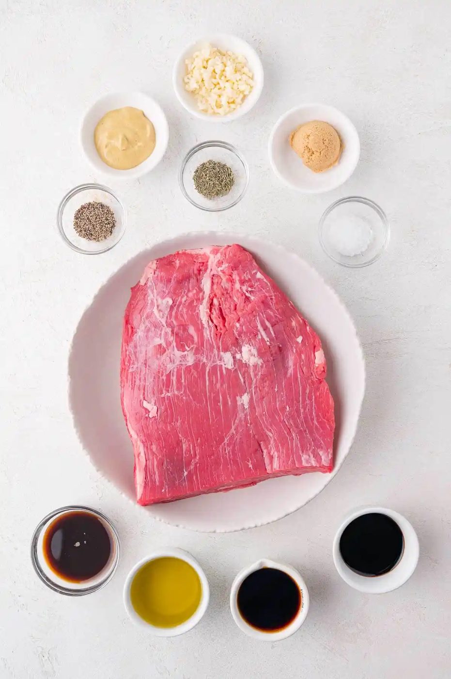 Raw flank steak on a plate surrounded by small bowls containing garlic, mustard, brown sugar, salt, pepper, dried herbs, soy sauce, olive oil, and Worcestershire sauce.