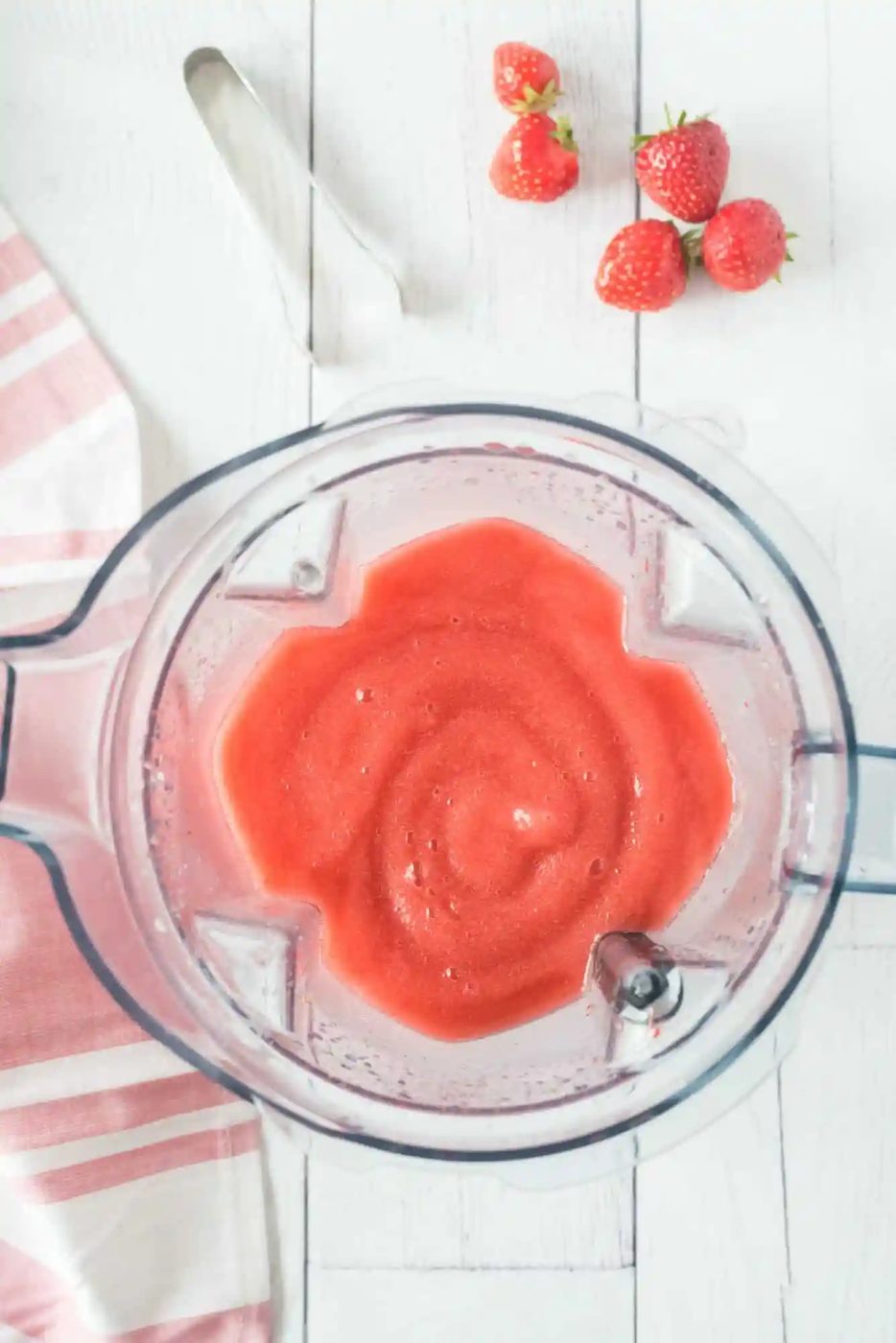 Blender with smooth frose on a white wooden surface, next to several whole strawberries and a pink-striped cloth.