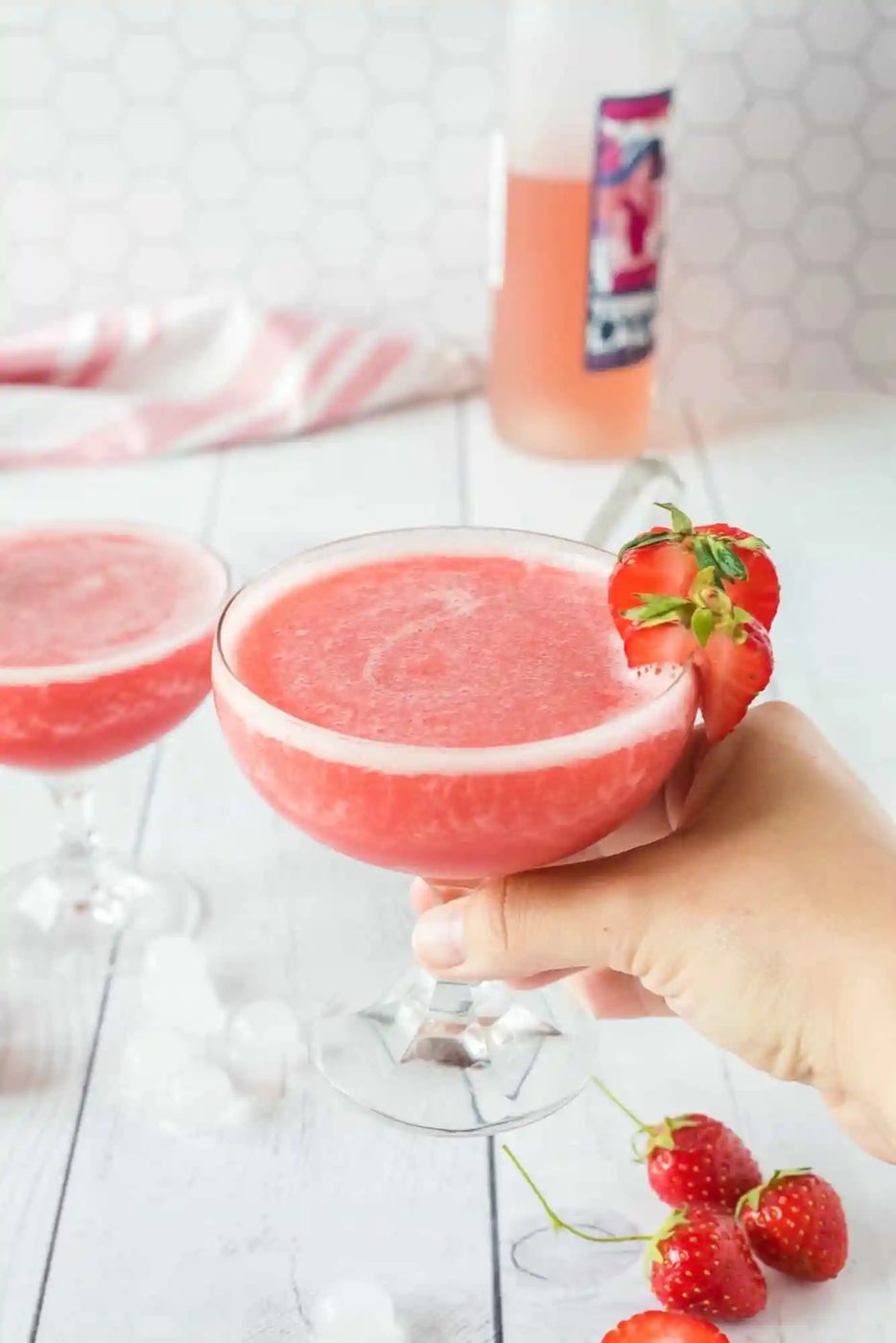 A hand holds a glass of pink cocktail garnished with a strawberry.