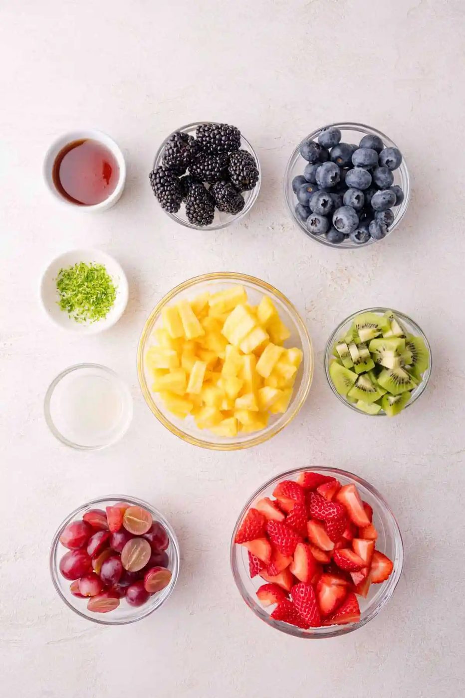 Bowls of cut pineapple, strawberries, grapes, kiwi, blackberries, and blueberries, with small dishes of lime juice, lime zest, and honey on a white surface.