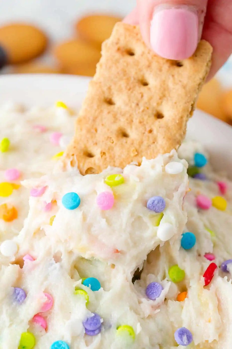 A hand dipping a graham cracker into a creamy dip topped with colorful sprinkles.