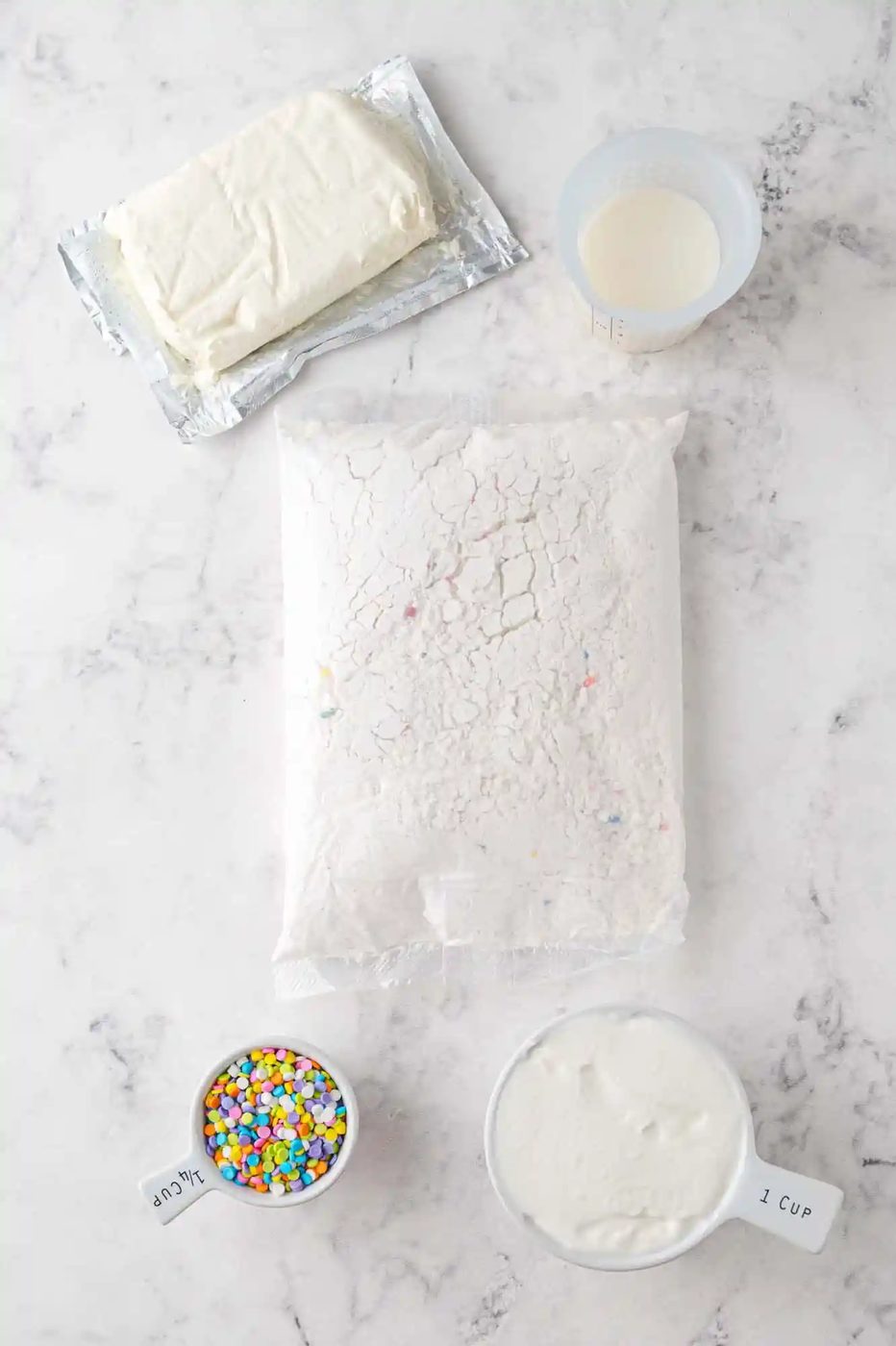 Ingredients on a marble surface: packaged cake mix, block of cream cheese, cup of milk, yogurt in a cup, and a bowl of colorful sprinkles.