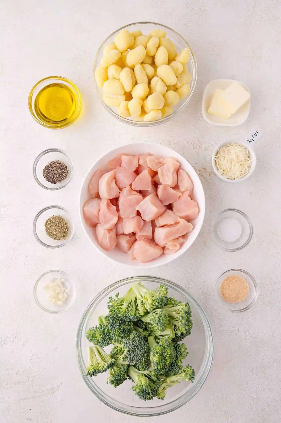 Top-down view of raw ingredients in bowls: gnocchi, chicken breast pieces, broccoli florets, olive oil, butter, grated cheese, minced garlic, and seasonings on a white surface.