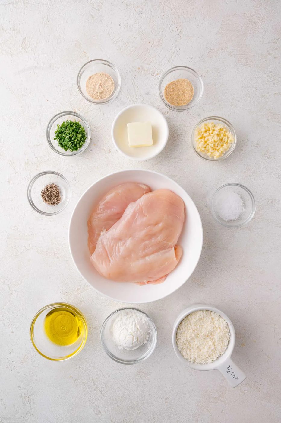 A bowl of raw chicken breasts surrounded by small bowls containing olive oil, butter, chopped herbs, garlic, grated cheese, and seasonings on a white surface.