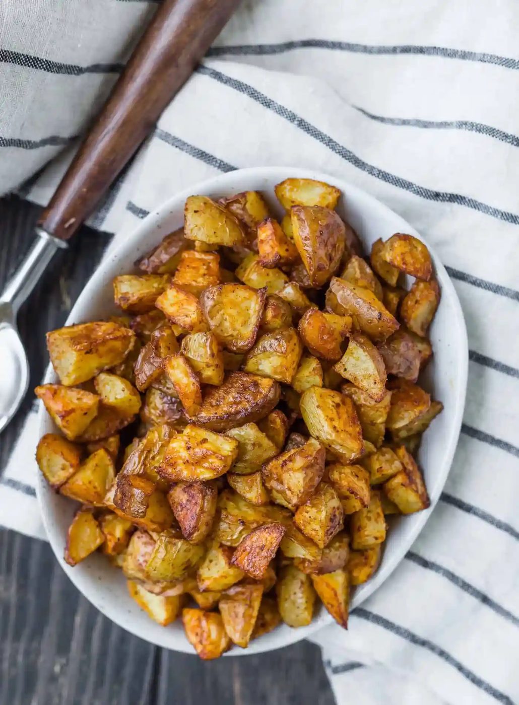 A white dish filled with roasted, diced potatoes sits on a striped cloth napkin, next to a metal serving spoon with a wooden handle.