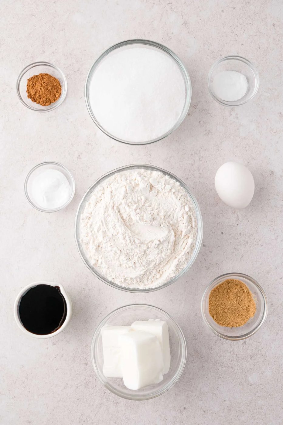 Bowls of flour, sugar, butter, molasses, eggs, baking soda, cinnamon, and salt arranged on a light surface.