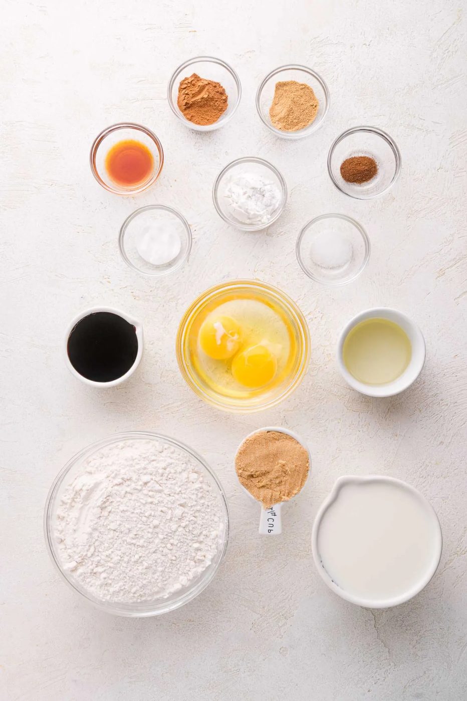 Overhead view of baking ingredients in bowls, including eggs, flour, sugar, spices, baking powder, oil, milk, molasses, vanilla extract, and brown sugar, arranged on a white surface.