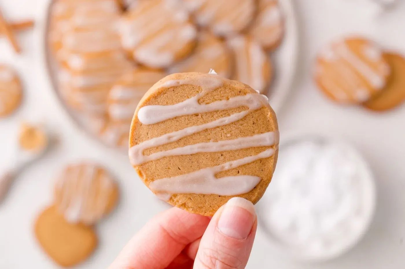 A hand holds a round gingerbread shortbread cookie with white icing drizzled on top; more cookies are visible blurred in the background.