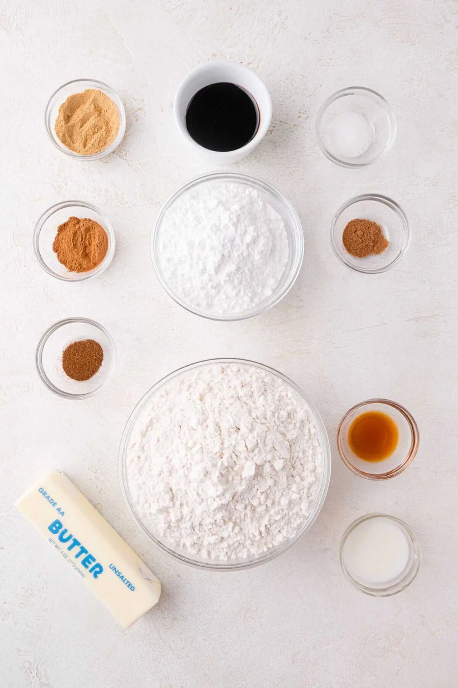 Overhead view of baking ingredients in small glass bowls, including flour, powdered sugar, ground spices, vanilla, milk, butter, and molasses, arranged on a white surface.