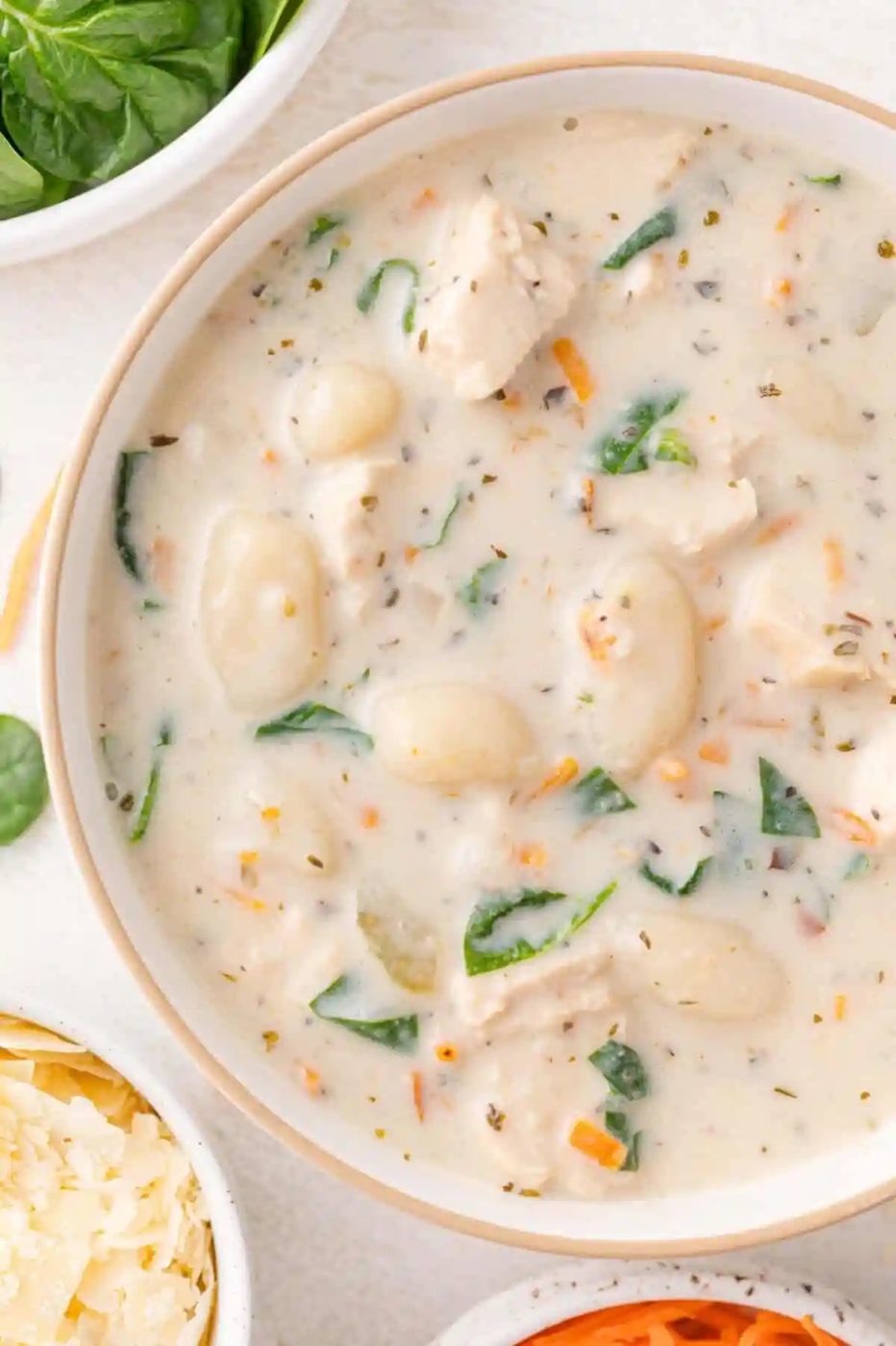 A bowl of creamy chicken gnocchi soup with spinach, carrots, and herbs, surrounded by bowls of grated cheese and fresh spinach leaves.