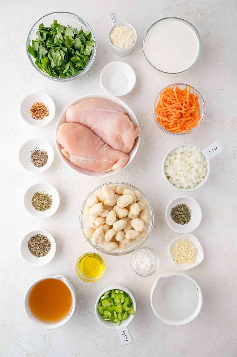 Ingredients for a chicken gnocchi soup recipe, including chicken breast, gnocchi, spinach, carrots, celery, chicken broth, milk, and various seasonings, arranged on a white surface.