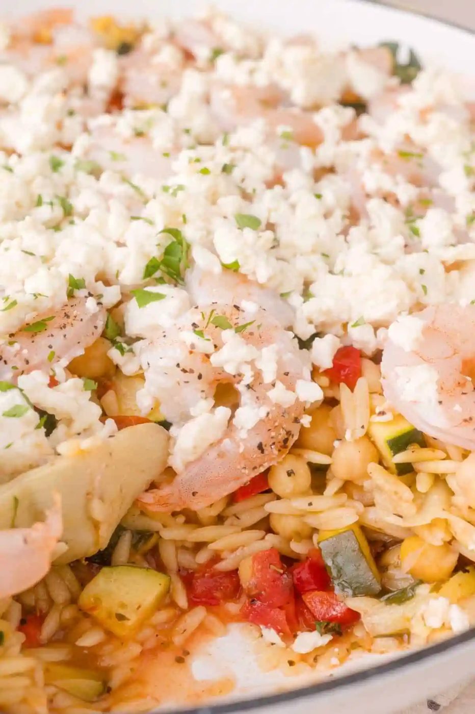 Close-up of a baked shrimp and vegetable orzo dish topped with crumbled feta cheese and garnished with herbs.