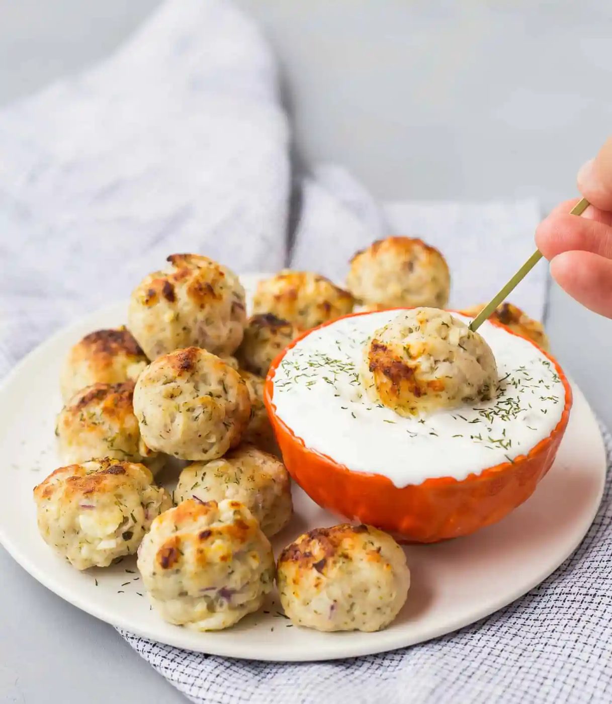 A plate of baked greek turkey meatballs is served with a bowl of white dipping sauce; one meatball is being dipped into the sauce using a skewer.