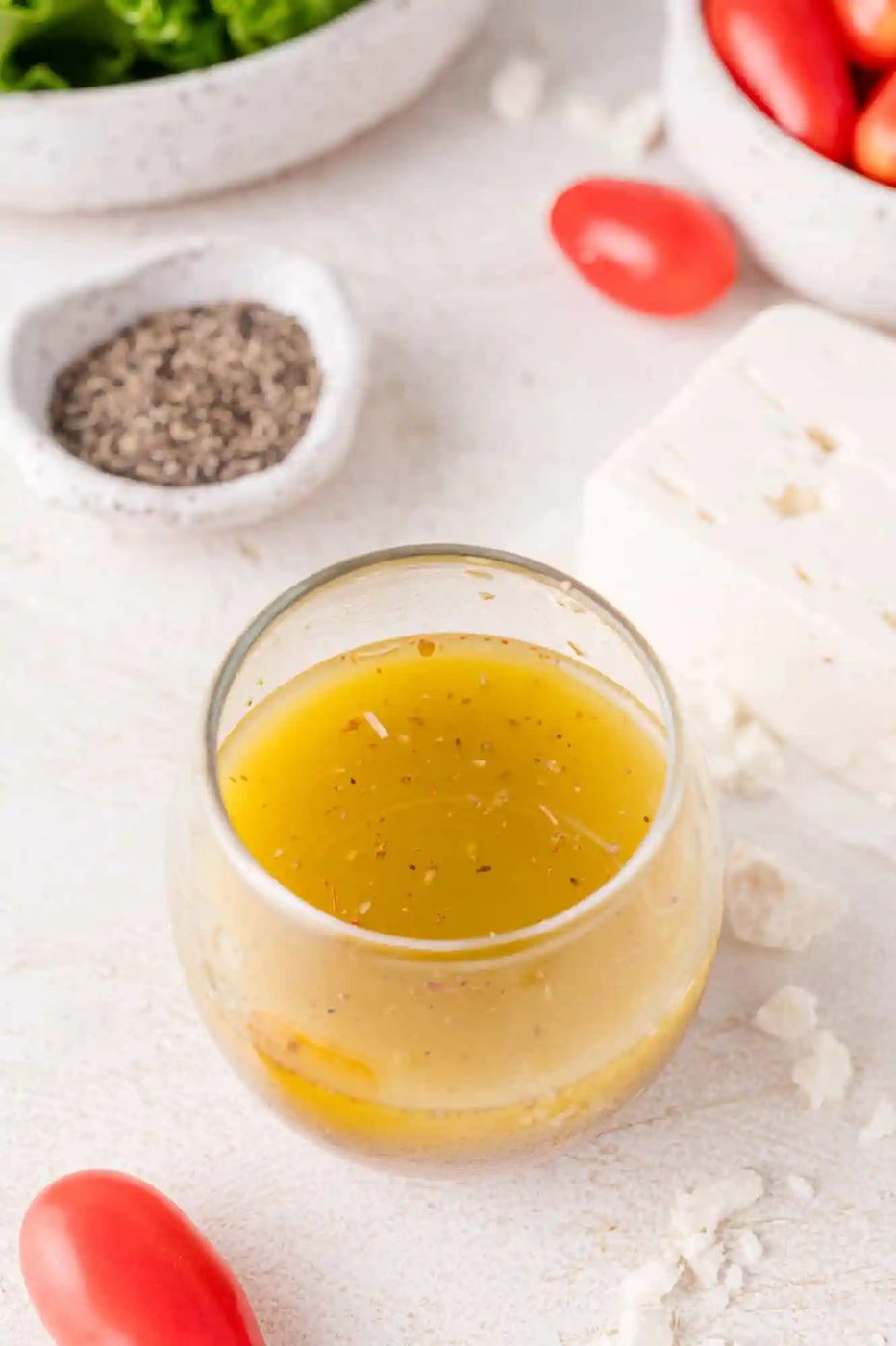 A glass bowl of salad dressing surrounded by pepper, feta cheese, cherry tomatoes, and lettuce on a white surface.