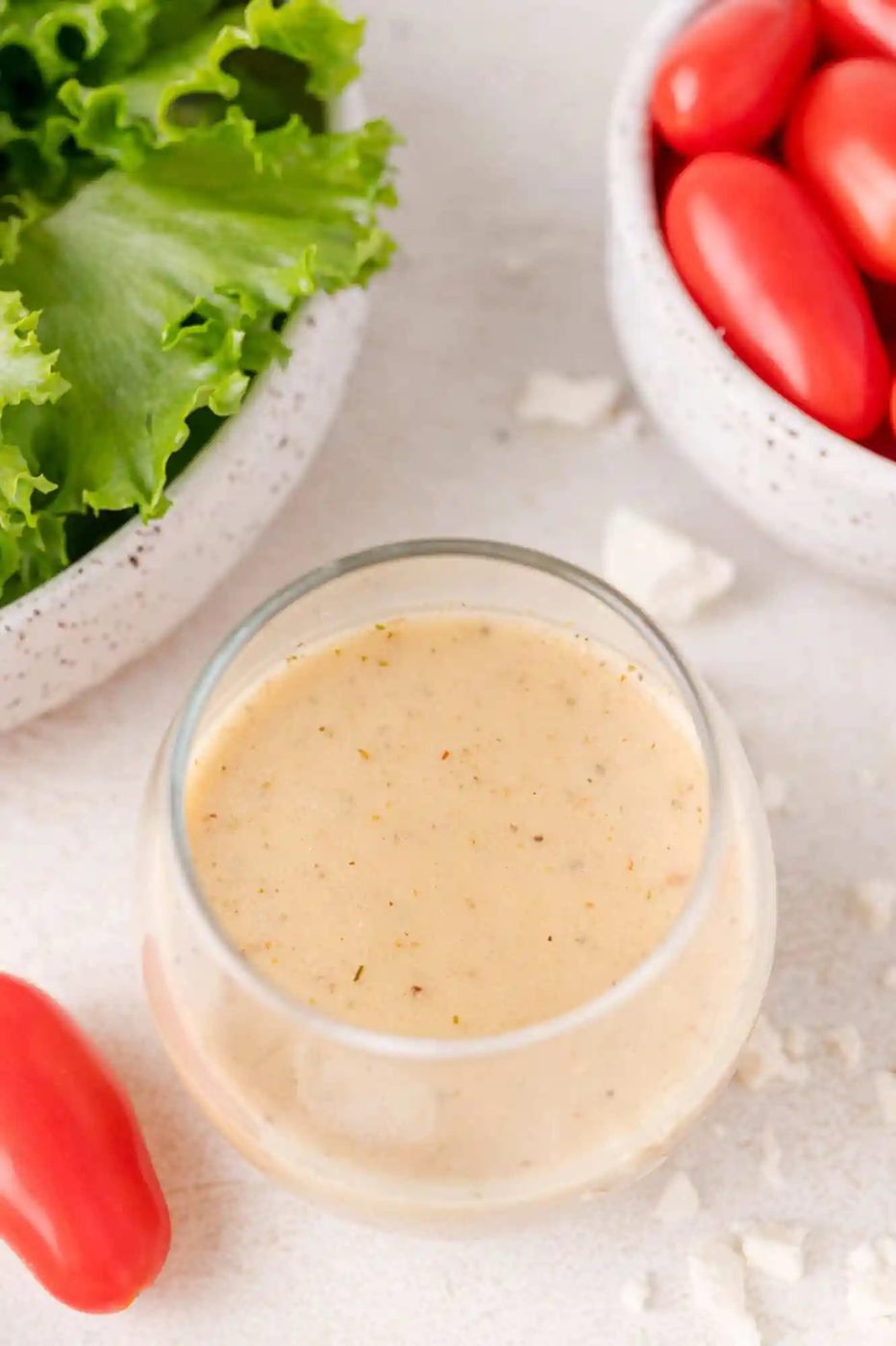 A glass bowl of creamy dressing, surrounded by a bowl of lettuce, cherry tomatoes, and crumbled cheese on a light surface.