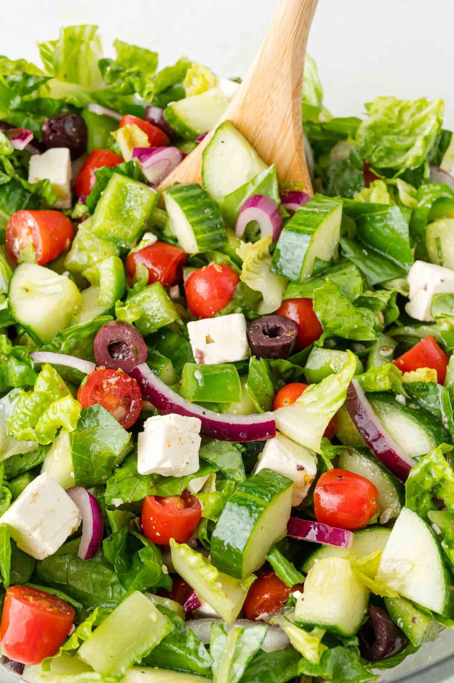 A salad with lettuce, cherry tomatoes, cucumbers, feta cheese, red onion, and olives, mixed with a wooden spoon.