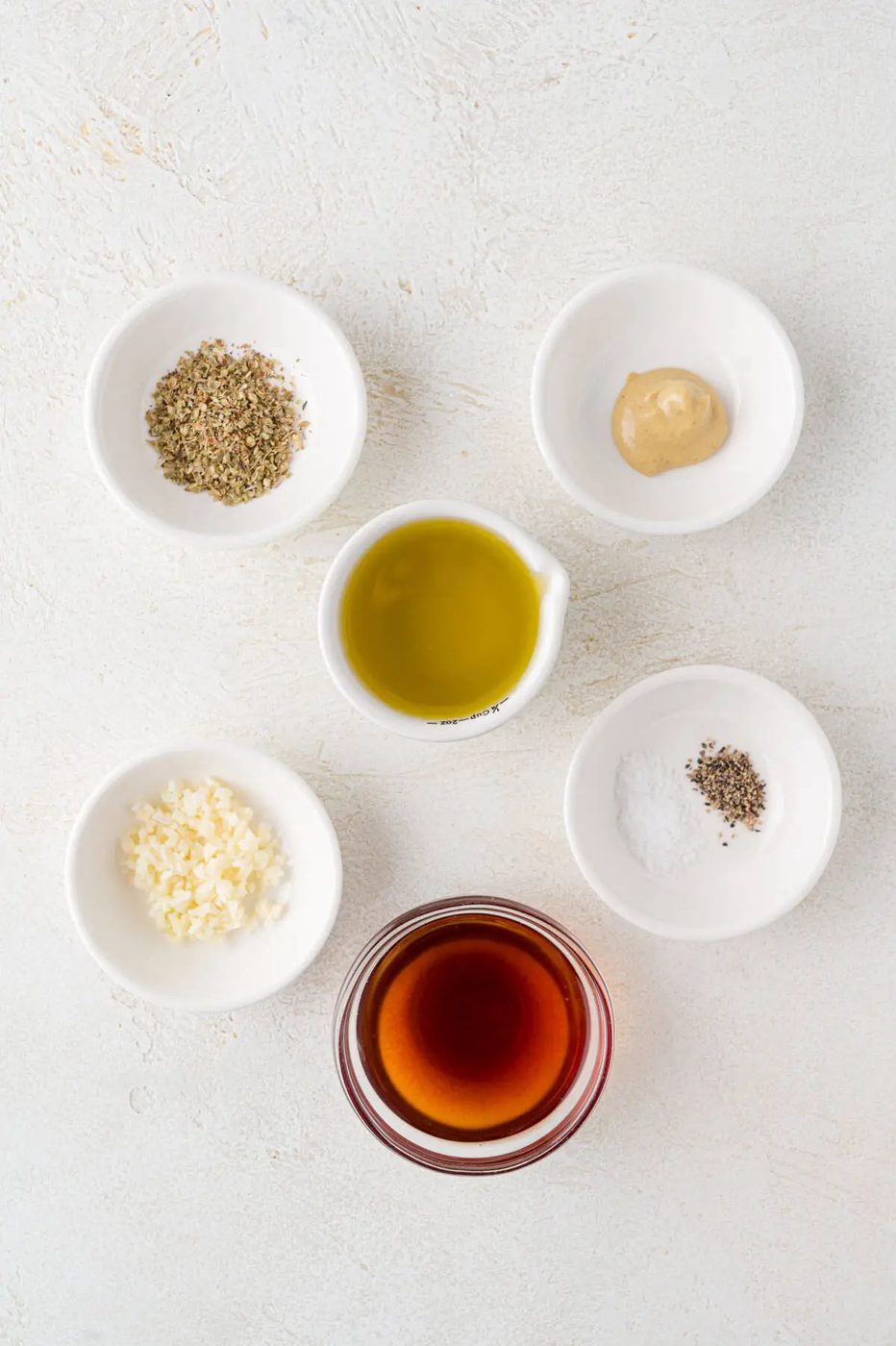 Five small bowls with salad dressing ingredients on a textured white surface.