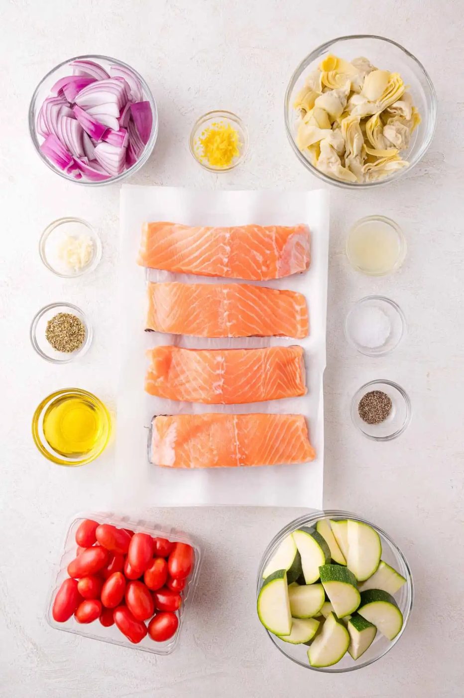 Overhead view of raw salmon fillets on parchment surrounded by bowls of red onion, artichoke hearts, zucchini, grape tomatoes, lemon zest, olive oil, seasoning, lemon juice, and garlic.