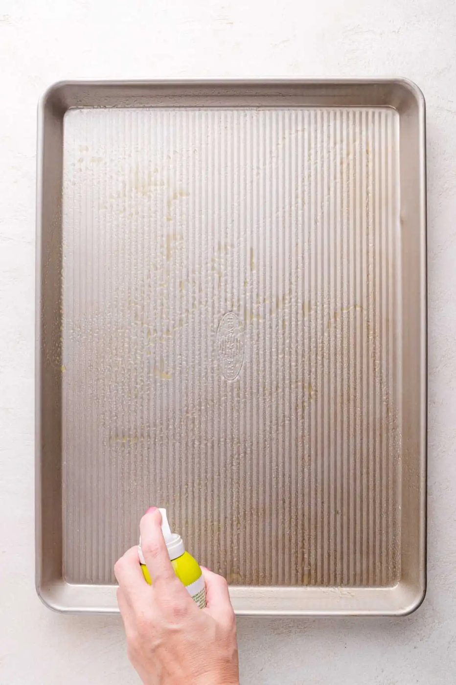 A hand sprays nonstick cooking spray onto a large, empty metal baking sheet placed on a light-colored surface.