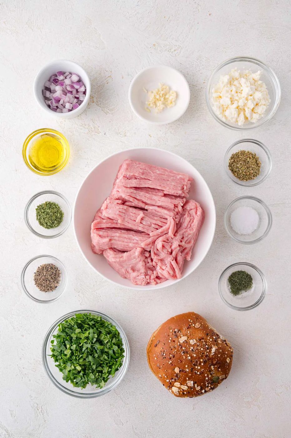 Ground turkey in a bowl surrounded by small bowls of chopped onion, garlic, parsley, feta, spices, olive oil, and an oat-topped burger bun on a white surface.