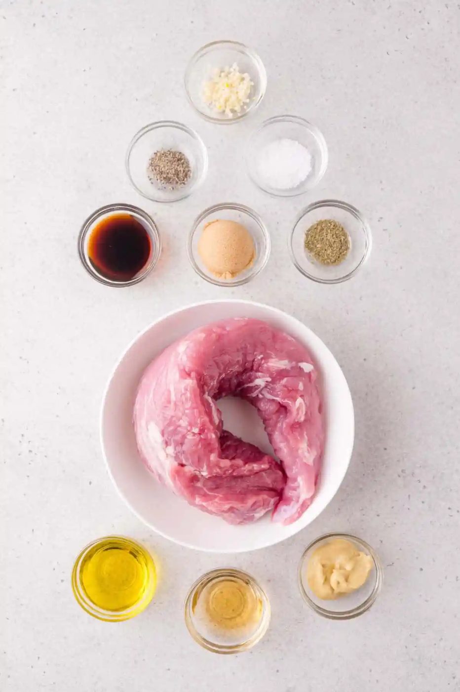 Raw pork tenderloin in a white bowl surrounded by small bowls of seasoning, oil, soy sauce, mustard, brown sugar, and minced garlic on a light surface.