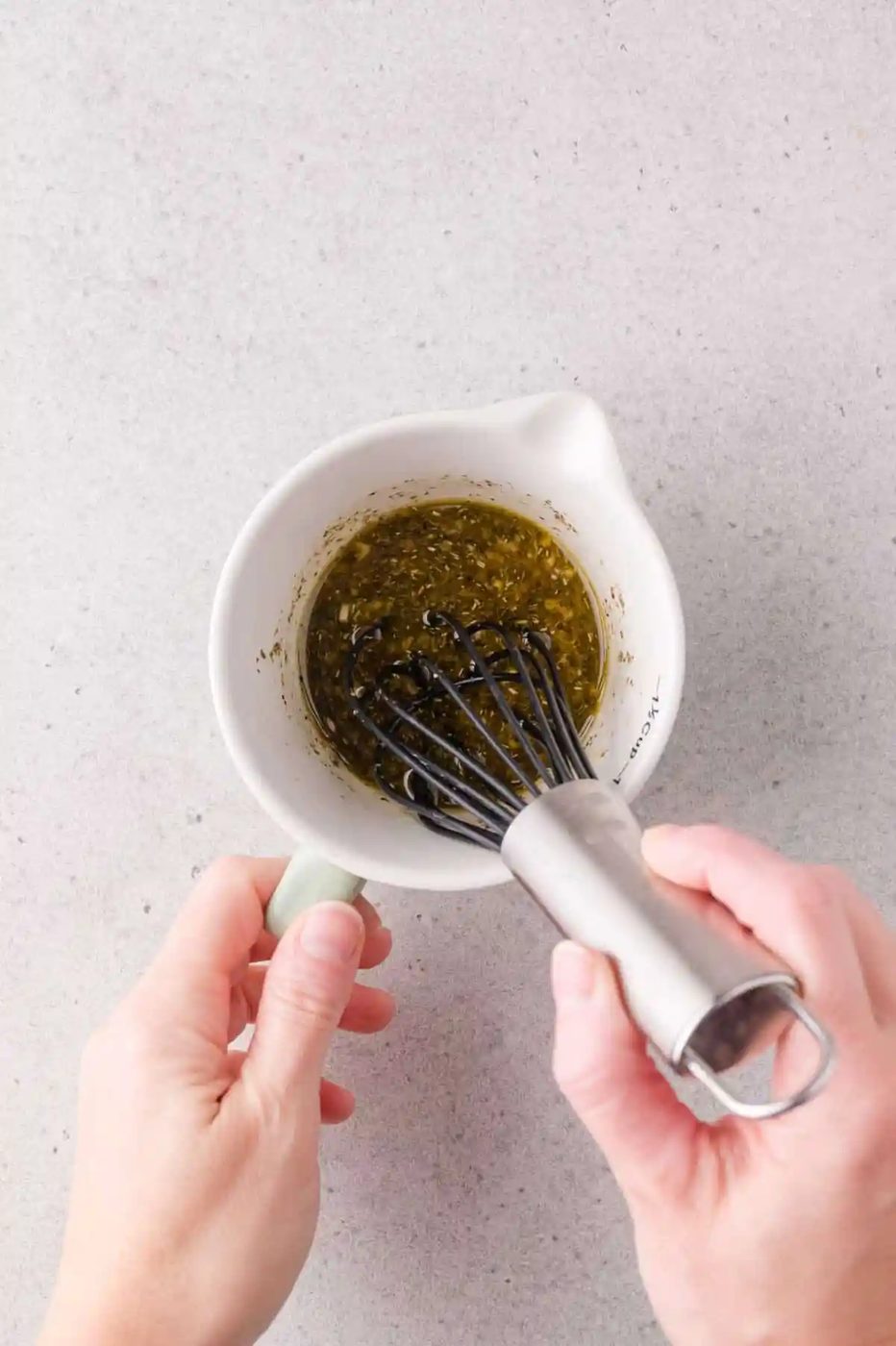 A person whisking marinade in a white ceramic measuring cup on a light gray surface.