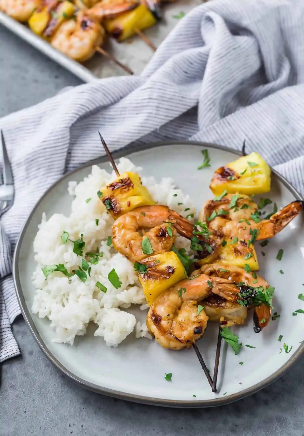 Two grilled shrimp kabobs with pineapple on a plate with fluffy white rice. More kabobs pictured in the background.