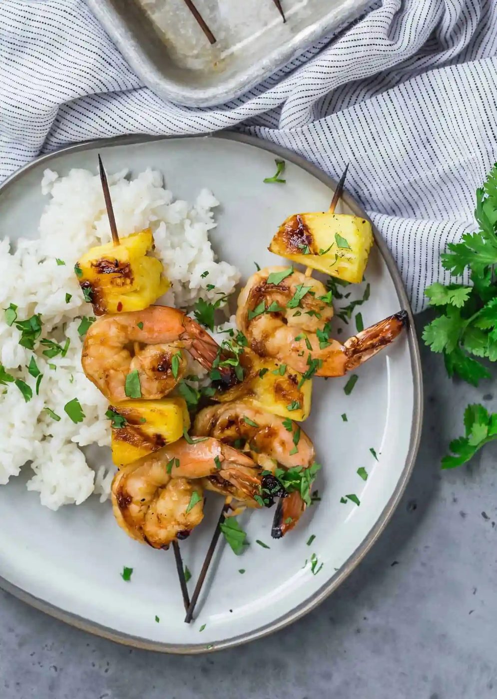 Overhead view of two grilled kabobs on a plate, with shrimp and fresh pineapple. They are served with white rice and sprinkled with fresh cilantro.