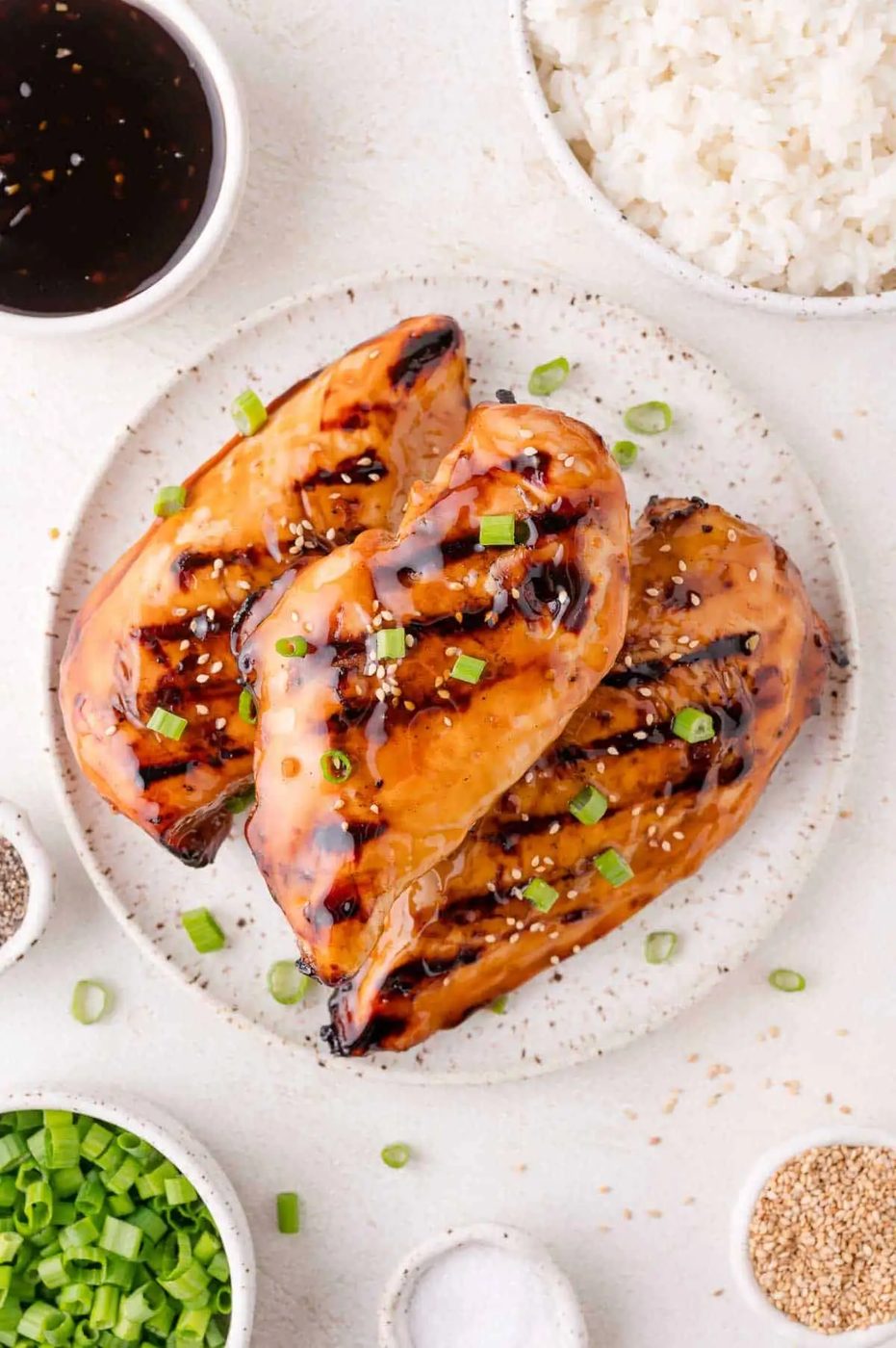Three grilled chicken breasts glazed with sauce, garnished with chopped green onions and sesame seeds, served on a plate with bowls of rice, sauce, green onions, pepper, and sesame seeds nearby.