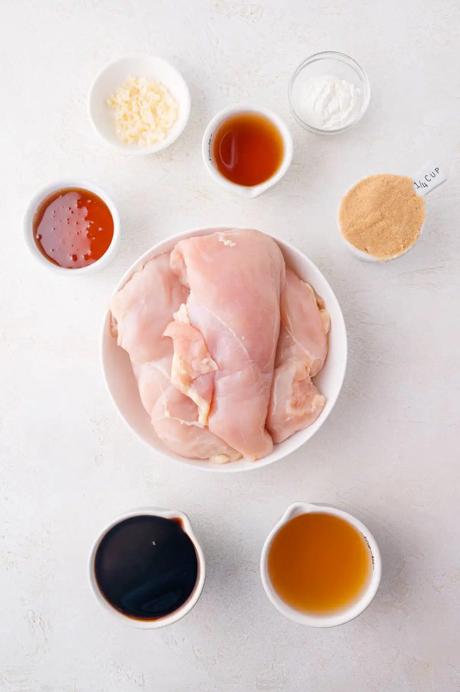 A bowl of raw chicken breasts surrounded by small bowls containing minced garlic, honey, soy sauce, brown sugar, apple cider vinegar, cornstarch, and another brown liquid on a white surface.